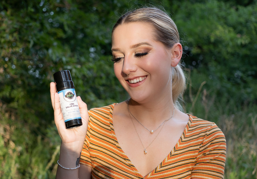 A young woman holding a cbd salve stick up in one hand while looking at it and smiling