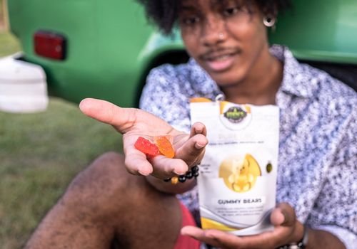 Young man sitting against a green van and holding handful of CBD gummies towards the camera