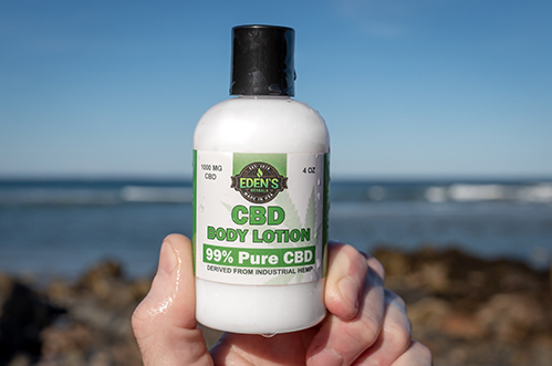 Hand holding up a bottle of Eden's Herbals CBD lotion with ocean in background