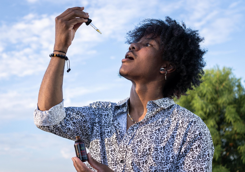 Young black man holding cbd oil tincture and placing dropper in mouth with clear blue sky behind him